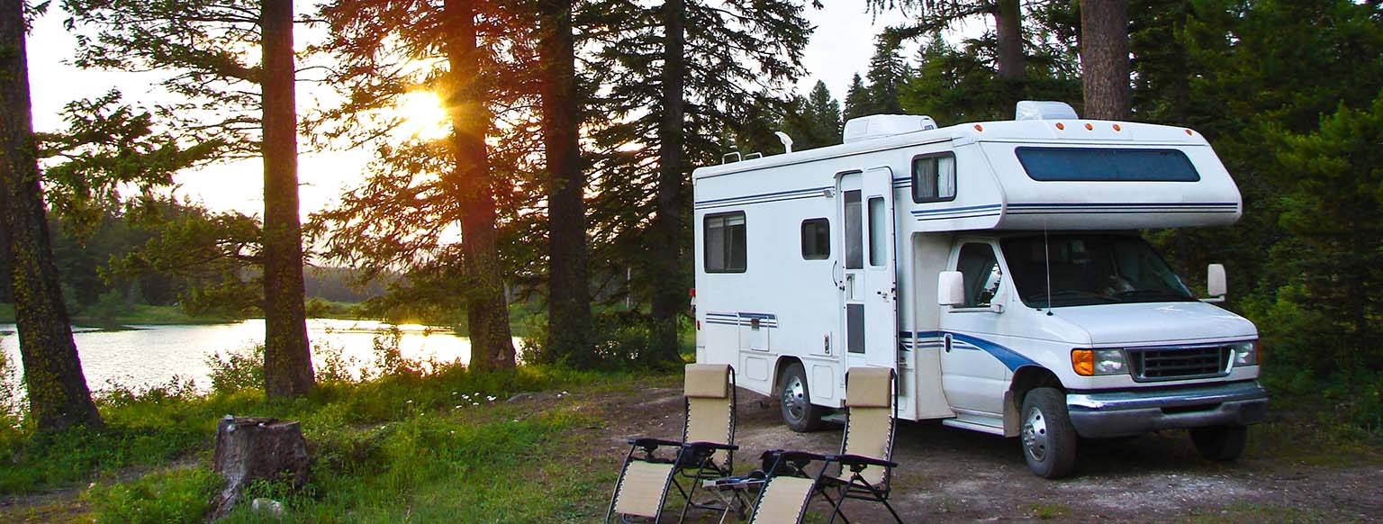 White camper parked in a forested area near a lake at sunset with two folding chairs - https://www.auburninsurance.org